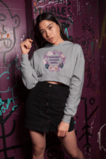 A young woman, possibly of East Asian descent, with dark hair, wearing a grey PodLyft female cropped hoodie. The hoodie features a circular graphic with floral elements and the empowering text "RESILIENT. RADIANT. RISING." in purple. She is posing casually against a graffiti-covered purple wall, looking towards the camera with a confident expression.