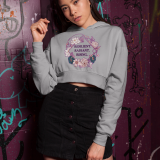 A young woman, possibly of East Asian descent, with dark hair, wearing a grey PodLyft female cropped hoodie. The hoodie features a circular graphic with floral elements and the empowering text "RESILIENT. RADIANT. RISING." in purple. She is posing casually against a graffiti-covered purple wall, looking towards the camera with a confident expression.