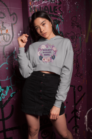 A young woman, possibly of East Asian descent, with dark hair, wearing a grey PodLyft female cropped hoodie. The hoodie features a circular graphic with floral elements and the empowering text "RESILIENT. RADIANT. RISING." in purple. She is posing casually against a graffiti-covered purple wall, looking towards the camera with a confident expression.
