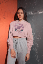 A young woman, possibly of East Asian descent, with dark hair, wearing a baby pink PodLyft female cropped hoodie. The hoodie displays a circular graphic with floral elements and the text "RESILIENT. RADIANT. RISING." in purple. She is standing with her hands in her pockets against a textured orange and grey wall, looking towards the camera.