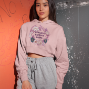 A young woman, possibly of East Asian descent, with dark hair, wearing a baby pink PodLyft female cropped hoodie. The hoodie displays a circular graphic with floral elements and the text "RESILIENT. RADIANT. RISING." in purple. She is standing with her hands in her pockets against a textured orange and grey wall, looking towards the camera.