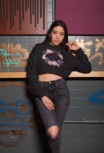 A young woman, possibly of East Asian descent, with dark hair, wearing a black PodLyft female cropped hoodie. The hoodie features a circular graphic with floral elements and the text "RESILIENT. RADIANT. RISING." in purple. She is leaning against a wooden surface in front of a tiled wall with graffiti, looking intently towards the camera.