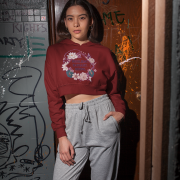 A young woman, possibly of East Asian descent, with dark hair, wearing a maroon PodLyft female cropped hoodie. The hoodie features a circular graphic with floral elements and the text "RESILIENT. RADIANT. RISING." in purple. She is standing with one hand in her pocket and the other near her face, against a graffiti-covered door in an urban setting.