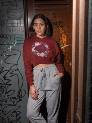 A young woman, possibly of East Asian descent, with dark hair, wearing a maroon PodLyft female cropped hoodie. The hoodie features a circular graphic with floral elements and the text "RESILIENT. RADIANT. RISING." in purple. She is standing with one hand in her pocket and the other near her face, against a graffiti-covered door in an urban setting.