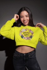 A young woman, possibly of East Asian descent, with dark hair, wearing a bright yellow PodLyft female cropped hoodie. The hoodie features a circular graphic with floral elements and the text "RESILIENT. RADIANT. RISING." in purple. She is smiling broadly and holding the hoodie near her shoulders, standing against a dark grey background.
