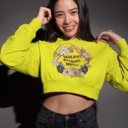 A young woman, possibly of East Asian descent, with dark hair, wearing a bright yellow PodLyft female cropped hoodie. The hoodie features a circular graphic with floral elements and the text "RESILIENT. RADIANT. RISING." in purple. She is smiling broadly and holding the hoodie near her shoulders, standing against a dark grey background.