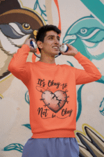 A young man, possibly of South Asian descent, wearing a coral-colored PodLyft men's mental health sweatshirt. The "It's Okay to Not Be Okay" design featuring a mended, cracked heart is visible. He is holding headphones to his ears and looking upwards, smiling, against a colorful mural background.