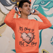 A young man, possibly of South Asian descent, wearing a coral-colored PodLyft men's mental health sweatshirt. The "It's Okay to Not Be Okay" design featuring a mended, cracked heart is visible. He is holding headphones to his ears and looking upwards, smiling, against a colorful mural background.