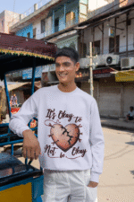 A young man, possibly of South Asian descent, wearing a lavender PodLyft men's mental health sweatshirt with the "It's Okay to Not Be Okay" design. The graphic features a mended, cracked heart. He is leaning on a rickshaw-like vehicle on a street that appears to be in India, looking to his left and smiling.