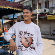 A young man, possibly of South Asian descent, wearing a lavender PodLyft men's mental health sweatshirt with the "It's Okay to Not Be Okay" design. The graphic features a mended, cracked heart. He is leaning on a rickshaw-like vehicle on a street that appears to be in India, looking to his left and smiling.
