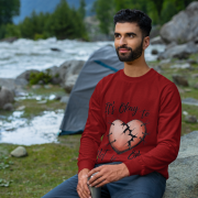 A man, possibly of Middle Eastern or South Asian descent, with dark curly hair and a beard, wearing a maroon PodLyft men's mental health sweatshirt. The sweatshirt displays the "It's Okay to Not Be Okay" design with a mended, cracked heart. He is sitting on a rock next to a flowing river, holding a silver water bottle, with mountains and trees in the background, looking into the distance.