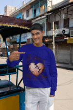 A young man, possibly of South Asian descent, wearing a navy blue PodLyft men's mental health sweatshirt. The "It's Okay to Not Be Okay" design with a mended, cracked heart is prominently displayed. He is standing next to a rickshaw-like vehicle on a street that appears to be in India, pointing to his chest and smiling at the camera.