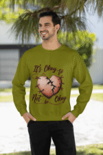 A man, possibly of Middle Eastern or South Asian descent, with dark hair and a beard, wearing an olive green PodLyft men's mental health sweatshirt. The sweatshirt features the "It's Okay to Not Be Okay" design with a mended, cracked heart. He is smiling and looking to his left, with his hands in his pockets, standing outdoors with trees and a building in the background.