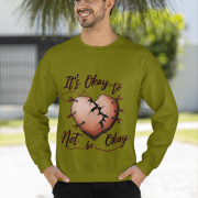 A man, possibly of Middle Eastern or South Asian descent, with dark hair and a beard, wearing an olive green PodLyft men's mental health sweatshirt. The sweatshirt features the "It's Okay to Not Be Okay" design with a mended, cracked heart. He is smiling and looking to his left, with his hands in his pockets, standing outdoors with trees and a building in the background.