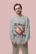 A man, possibly of Caucasian descent, with a beard and medium-length hair, wearing a light grey PodLyft's mental health sweatshirt. The sweatshirt features a graphic of a pink heart with cracks mended by red stitching and pins, with the text "It's Okay to Not be Okay" in red script above and below the heart. He is looking directly at the camera with a serious expression, standing against a plain light pink background.