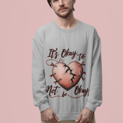 A man, possibly of Caucasian descent, with a beard and medium-length hair, wearing a light grey PodLyft's mental health sweatshirt. The sweatshirt features a graphic of a pink heart with cracks mended by red stitching and pins, with the text "It's Okay to Not be Okay" in red script above and below the heart. He is looking directly at the camera with a serious expression, standing against a plain light pink background.