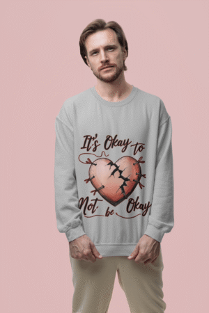 A man, possibly of Caucasian descent, with a beard and medium-length hair, wearing a light grey PodLyft's mental health sweatshirt. The sweatshirt features a graphic of a pink heart with cracks mended by red stitching and pins, with the text "It's Okay to Not be Okay" in red script above and below the heart. He is looking directly at the camera with a serious expression, standing against a plain light pink background.