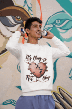 A young man, possibly of South Asian descent, wearing a white PodLyft men's mental health sweatshirt with the "It's Okay to Not Be Okay" design. The graphic features a mended, cracked heart. He is holding headphones to his ears and looking upwards, smiling, against a colorful mural background.