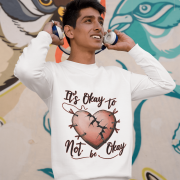 A young man, possibly of South Asian descent, wearing a white PodLyft men's mental health sweatshirt with the "It's Okay to Not Be Okay" design. The graphic features a mended, cracked heart. He is holding headphones to his ears and looking upwards, smiling, against a colorful mural background.