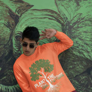 A man wearing a coral PodLyft Men's Plant the Future Sweatshirt, featuring a prominent tree design with roots and the text "PLANT THE FUTURE," posing in front of a green mural.
