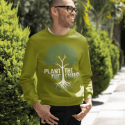 A smiling man wearing an olive green PodLyft Men's Plant the Future Sweatshirt, featuring a prominent tree design with roots and the text "PLANT THE FUTURE," standing outdoors with greenery in the