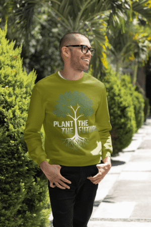 A smiling man wearing an olive green PodLyft Men's Plant the Future Sweatshirt, featuring a prominent tree design with roots and the text "PLANT THE FUTURE," standing outdoors with greenery in the