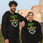 A man and a woman, possibly a couple of South Asian descent, standing outdoors in front of a light-colored historical building. Both are wearing black Unity in Diversity hoodies with a graphic of hands of various skin tones forming a heart shape. The man, with curly dark hair, has his arm resting on the woman's shoulder.