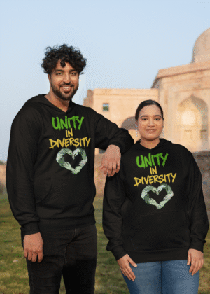 A man and a woman, possibly a couple of South Asian descent, standing outdoors in front of a light-colored historical building. Both are wearing black Unity in Diversity hoodies with a graphic of hands of various skin tones forming a heart shape. The man, with curly dark hair, has his arm resting on the woman's shoulder.