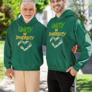 Two men, one older and one younger, standing side-by-side outdoors with green trees and a building in the background. Both are smiling and wearing bottle green Unity in Diversity hoodies. The hoodies feature a graphic of hands of various skin tones forming a heart shape. The younger man has his arm around the older man's back.