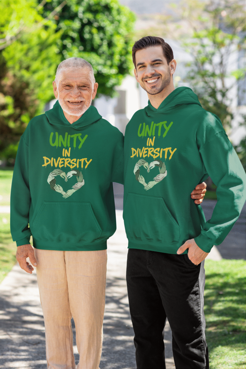 Two men, one older and one younger, standing side-by-side outdoors with green trees and a building in the background. Both are smiling and wearing bottle green Unity in Diversity hoodies. The hoodies feature a graphic of hands of various skin tones forming a heart shape. The younger man has his arm around the older man's back.