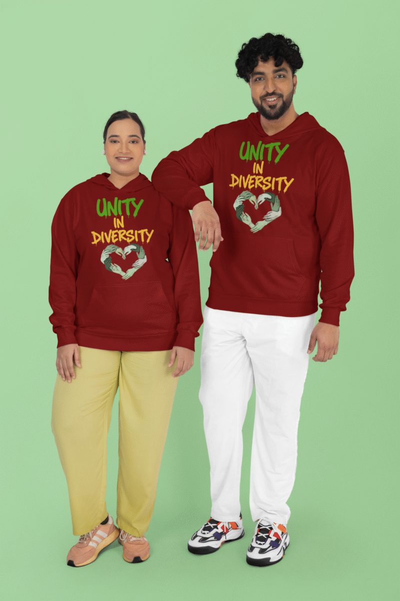A man and a woman, possibly a couple of South Asian descent, standing in front of a light green background. Both are wearing maroon Unity in Diversity hoodies with a graphic of hands of various skin tones forming a heart shape. The man, with curly dark hair, has his arm resting on the woman's shoulder.