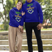 A man and a woman, possibly a couple, standing on outdoor steps with green foliage in the background. They are both wearing navy blue Unity in Diversity hoodies, which feature a graphic of hands of various skin tones forming a heart shape. The woman, with dark hair, leans her head on the man's shoulder. The man, with short curly hair, smiles.