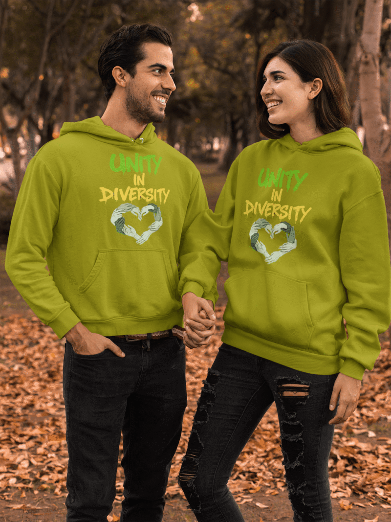 A man and a woman, possibly a couple, holding hands and looking at each other in an outdoor setting with autumn leaves on the ground and blurred trees in the background. They are both wearing olive green Unity in Diversity hoodies, which feature a graphic of hands of various skin tones forming a heart shape.