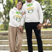 A man and a woman, possibly a couple, standing on outdoor steps with green foliage in the background. They are both wearing white Unity in Diversity hoodies. The graphic features hands of various skin tones forming a heart shape. The woman, with dark hair, leans her head on the man's shoulder. The man, with short curly hair, smiles.