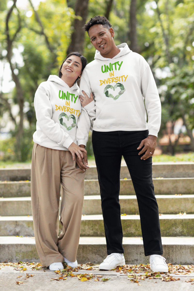 A man and a woman, possibly a couple, standing on outdoor steps with green foliage in the background. They are both wearing white Unity in Diversity hoodies. The graphic features hands of various skin tones forming a heart shape. The woman, with dark hair, leans her head on the man's shoulder. The man, with short curly hair, smiles.