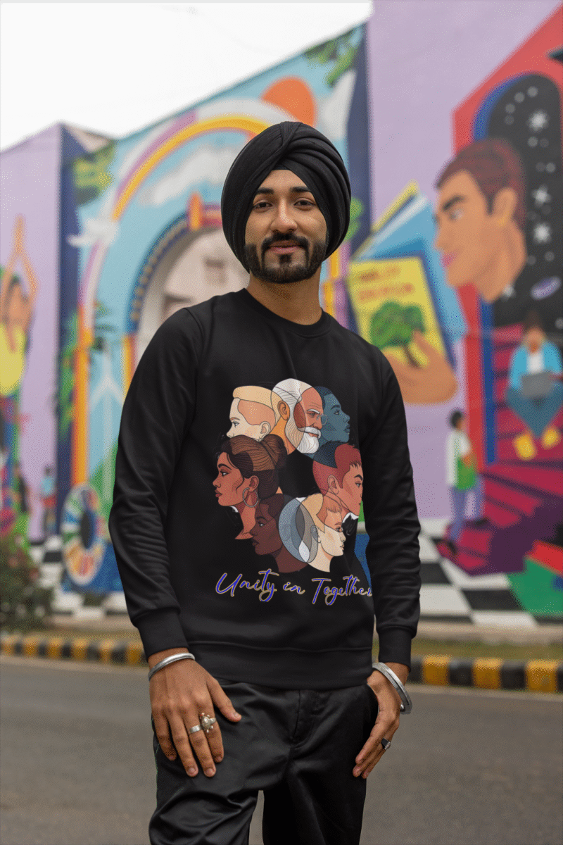A man, possibly of South Asian descent, wearing a black turban and a black Unity in Together sweatshirt. The sweatshirt features a circular graphic with diverse profiles of people. He is standing outdoors in front of a colorful mural, looking towards the camera and smiling. He is wearing metal bracelets and rings.