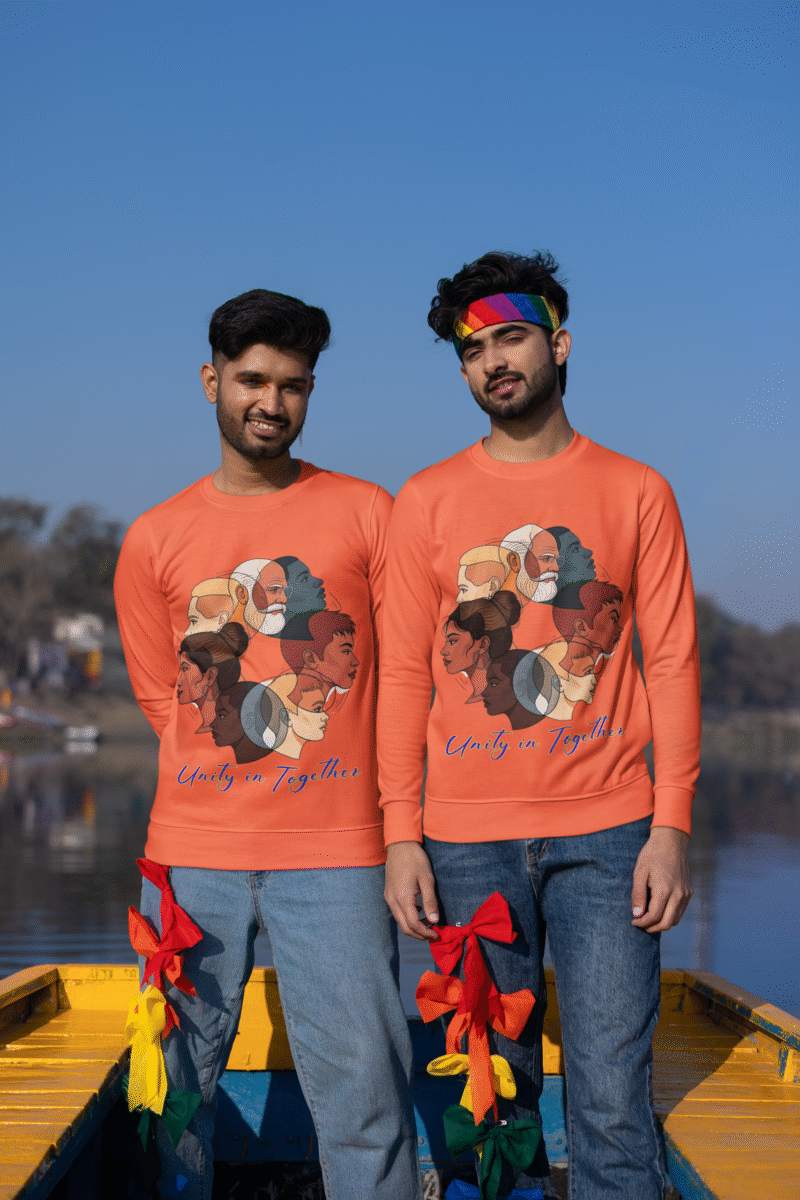 Two young men, possibly of South Asian descent, standing in a boat on a body of water under a clear blue sky. Both are wearing coral-colored Unity in Together sweatshirts that feature a circular graphic with diverse profiles of people. One man is wearing a rainbow-colored headband and both have festive red and yellow ribbons tied to their jeans.