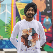 A man, possibly of South Asian descent, wearing a black turban and a white Unity in Together sweatshirt. The sweatshirt features a circular graphic with diverse profiles of people. He is standing outdoors in front of a vibrant, colorful mural, looking towards the camera with a gentle smile.