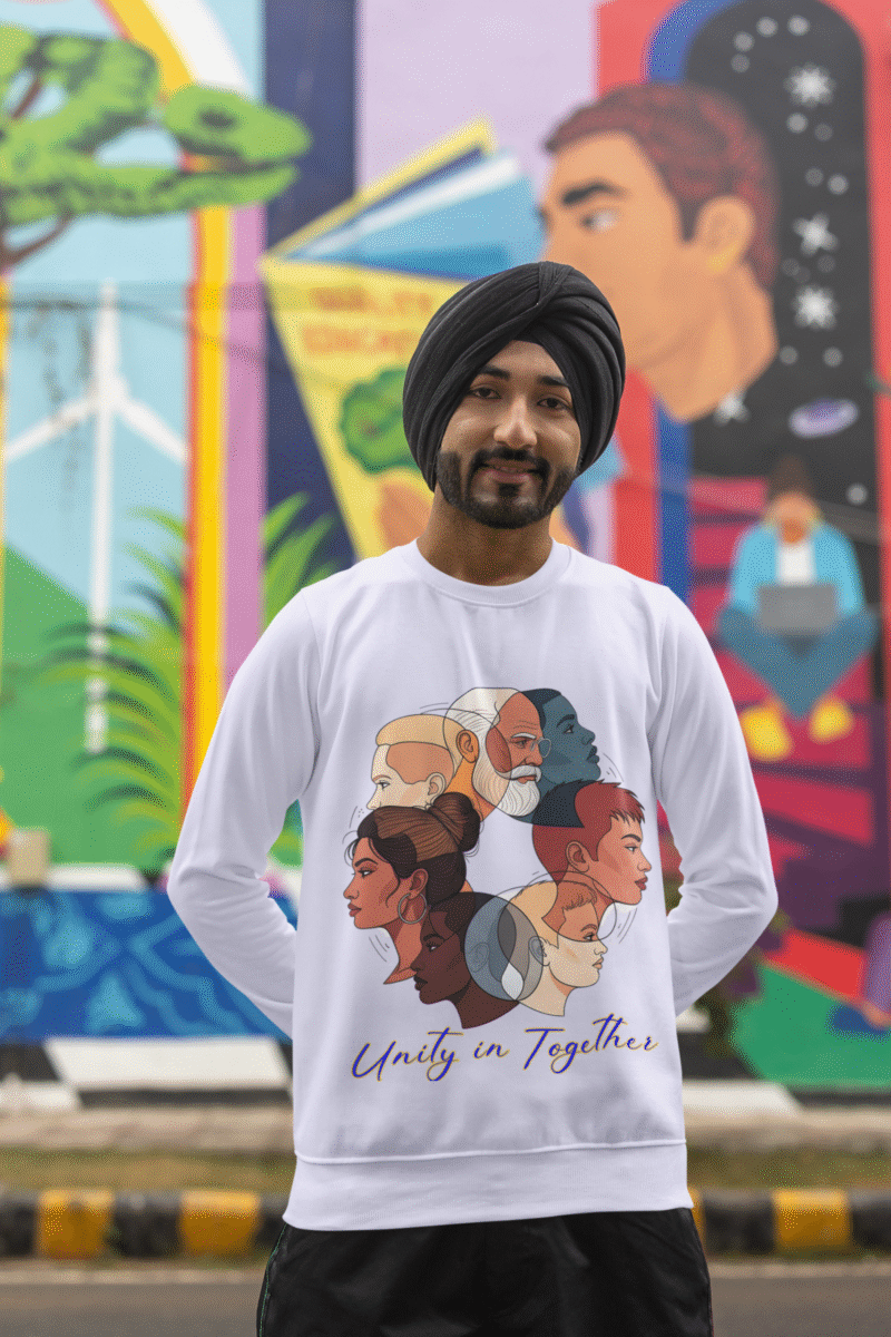 A man, possibly of South Asian descent, wearing a black turban and a white Unity in Together sweatshirt. The sweatshirt features a circular graphic with diverse profiles of people. He is standing outdoors in front of a vibrant, colorful mural, looking towards the camera with a gentle smile.