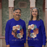 An older couple, possibly of South Asian descent, smiling and standing side-by-side in front of an architectural backdrop with arched windows. Both are wearing navy blue Unity in Together sweatshirts, which feature a circular graphic with diverse profiles of people. The man has a mustache and is wearing a dark lungi-style garment, and the woman has long grey hair.