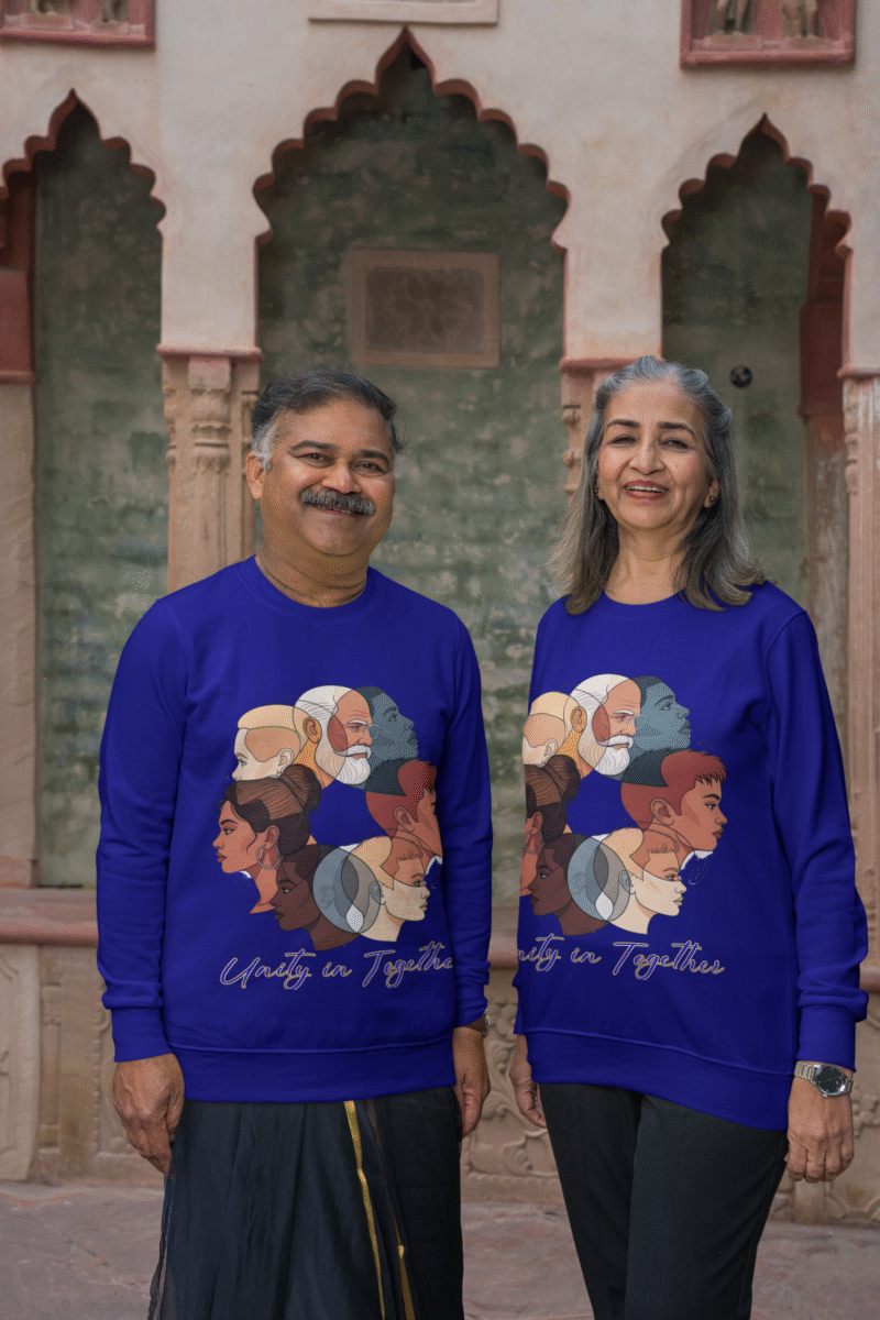 An older couple, possibly of South Asian descent, smiling and standing side-by-side in front of an architectural backdrop with arched windows. Both are wearing navy blue Unity in Together sweatshirts, which feature a circular graphic with diverse profiles of people. The man has a mustache and is wearing a dark lungi-style garment, and the woman has long grey hair.