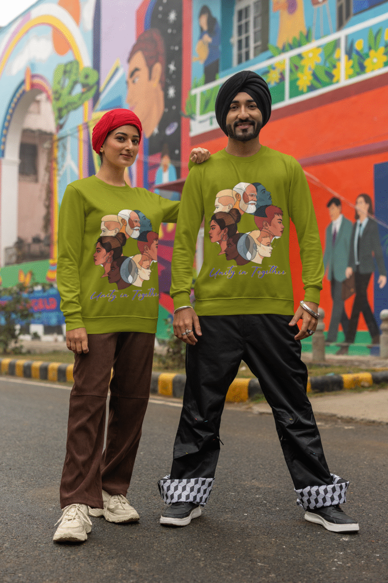 A couple, possibly of South Asian descent, standing side-by-side in front of a colorful mural. The man, wearing a black turban, and the woman, wearing a red headscarf, are both wearing olive green Unity in Together sweatshirts. The sweatshirts feature a circular graphic with diverse profiles of people. The woman has her arm around the man's shoulder.