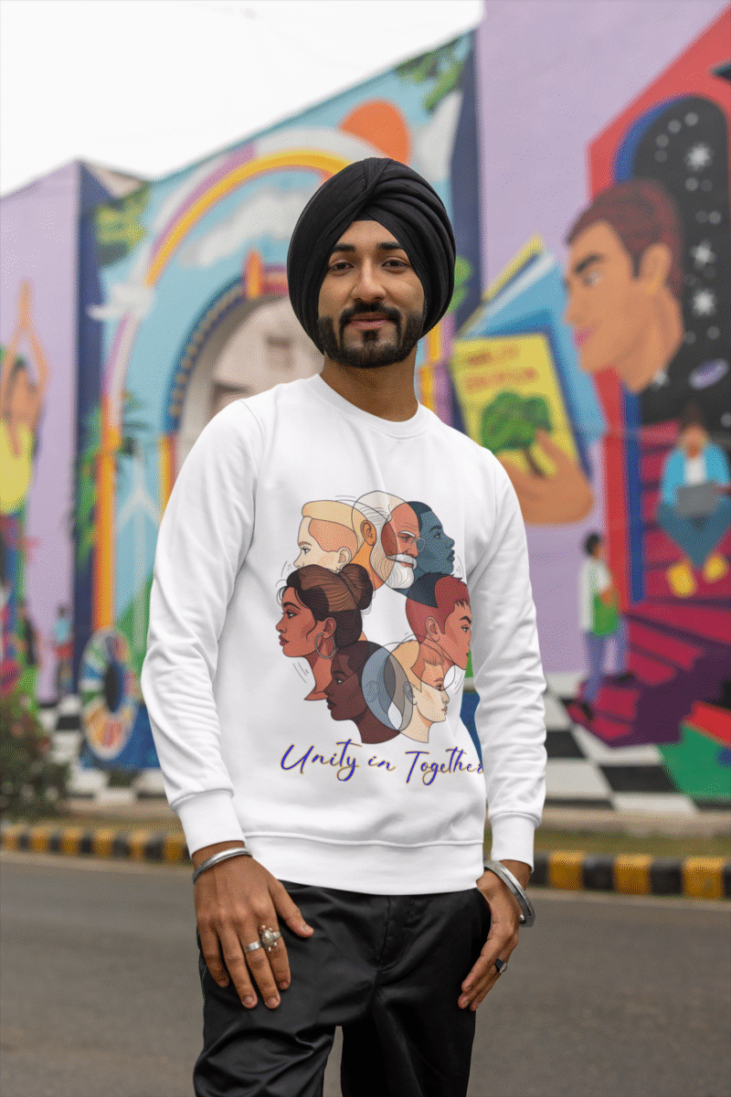 A man, possibly of South Asian descent, wearing a black turban and a white "Unity in Together" sweatshirt. The sweatshirt features a circular graphic with diverse profiles of people. He is standing outdoors in front of a colorful mural, looking towards the camera and smiling. He is wearing metal bracelets and rings.