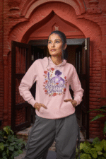 A young woman, possibly of East Asian descent, with dark hair, wearing a baby pink PodLyft women's empowered hoodie. The hoodie features an abstract graphic with geometric shapes and subtle lines in shades of purple and pink, conveying a message of empowerment. She is smiling confidently, with her hands in her pockets, standing against a blurred grey outdoor background.
