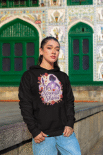 A young woman, possibly of East Asian descent, with dark hair, wearing a black PodLyft women's empowered hoodie. The hoodie features an abstract graphic with geometric shapes and subtle lines in shades of purple and pink, conveying a message of empowerment. She is looking directly at the camera with a confident expression, standing against a blurred grey outdoor background.