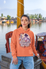A young woman, possibly of East Asian descent, with dark hair, wearing a coral PodLyft women's empowered hoodie. The hoodie features an abstract graphic with geometric shapes and subtle lines in shades of purple and pink, conveying a message of empowerment. She is smiling confidently, with her hands in her pockets, standing against a blurred outdoor background.