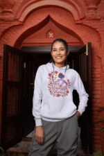 A young woman, possibly of East Asian descent, with dark hair, wearing a lavender PodLyft women's empowered hoodie. The hoodie features an abstract graphic with geometric shapes and subtle lines in shades of purple and pink, conveying a message of empowerment. She is looking directly at the camera with a confident expression, standing against a blurred grey background.