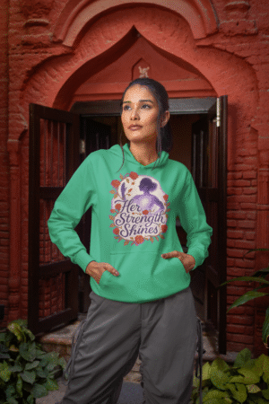 A young woman, possibly of East Asian descent, with dark hair, wearing a mint green PodLyft women's empowered hoodie. The hoodie features an abstract graphic with geometric shapes and subtle lines in shades of purple and pink, conveying a message of empowerment. She is smiling confidently, holding a small purple clutch bag, standing against a blurred outdoor background.