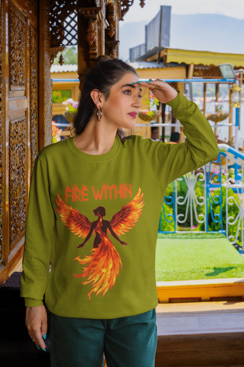 A young woman, possibly of Caucasian descent with light brown hair, wearing an olive green PodLyft women's Fire Within sweatshirt. The sweatshirt features a graphic of a human heart with stylized flames emanating from its top, and the text "FIRE WITHIN" written above and below it. She is looking directly at the camera with a subtle smile, standing against a plain light grey background.