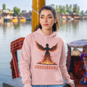 A young woman, possibly of Caucasian descent, with dark hair tied back, wearing a baby pink PodLyft women's Unbreakable Spirit hoodie. The hoodie features a graphic of a stylized, strong female figure in motion, with the word "UNBREAKABLE" across her. She is looking directly at the camera with a determined expression, standing against a plain light grey background.